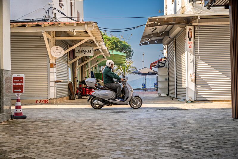 A scooter crosses the end of the street, the turquoise boat sits in the marina, and a couple walk down towards the water. (1 of 3)